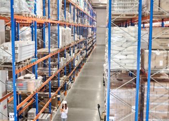 Large warehouse. Woman in white protective helmet with tablet standing in large industrial warehouse in aisle between high racks for goods